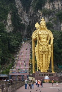 Golden Lord Murugan statue next to stairs leading into cave.